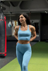 Fată brunetă purtând set sport GYMTUNED albastru pastel – bustieră și colanți, poză în sală de fitness cu sac de box în fundal